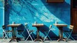 Four wooden tables with matching folding chairs are set up on a sidewalk against a vibrant blue wall, with the morning sunlight casting shadows of tree branches on the painted surface, creating a sere