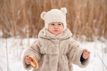 Wall Mural - A cute little kid in a plush hat and a fur coat has a steering wheel on the background of a snowy winter landscape with reeds. The winter season, a cozy childhood. Russian Slavic style.