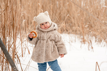 Wall Mural - A cute little kid in a plush hat and a fur coat eats a bagel on the background of a snowy winter landscape with reeds. The winter season, a cozy childhood. Russian Slavic style.