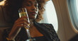 Lifestyle portrait of attractive wealthy black woman passenger seated in window seat on private jet and drinking glass of champagne on flight