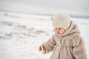 Wall Mural - Cute little kid in a plush hat and fur coat eats a bagel on the background of a snowy winter landscape on the river,white wasteland