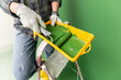 Person on a stepledder  in working gloves holding yellow paint trough with middle-sized paint roller with green paint in front of  green wall.