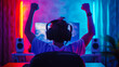 © Davin - Excited gamer with headphones raising fists in triumph in a vibrant, neon-lit gaming setup at night. E-sport technology content creator lifestyle concept.