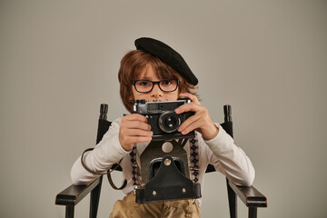 Wall Mural - boy in beret and glasses holding vintage camera while sitting on director chair, young photographer