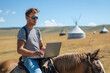 A young digital nomad wearing sunglasses sits on a horse with his laptop in a field, traditional yurts visible in the background under a clear blue sky.