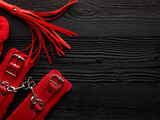 Fototapeta  - Bright red handcuffs and red whip over black wooden background