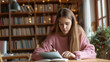 © Marta B - a young woman is studying intently in a library, writing in a notebook while looking at a laptop screen, surrounded by bookshelves.