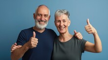älteres Paar Zeigt Daumen Hoch – Zustimmung, Aufbruch, Optimismus. Sie Tragen Dunkle T-Shirts Vor Einer Hellblauen Wand.