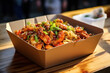Asian dish noodles with chicken and vegetables in cardboard box on table in street eatery, concept fast to go street food