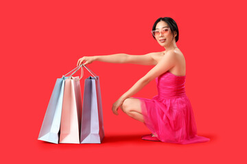 Sticker - Happy beautiful young Asian woman with shopping bags sitting on red background