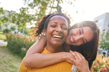 Wall Mural - Portrait young friends happy in park flare lens in background. Attractive Caucasian woman hugs smiling latina girl in affectionate attitude. Multiracial females looking at cheerful camera outdoor.