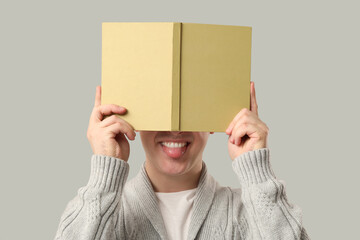 Wall Mural - Young man covering his face with book and showing tongue on grey background, closeup