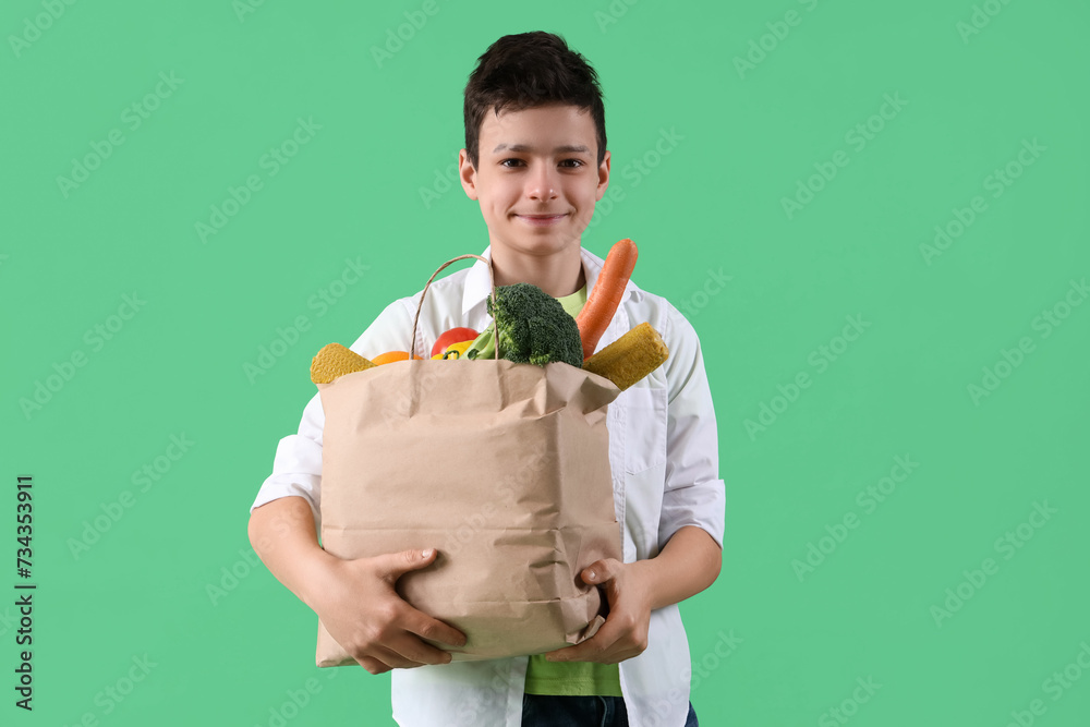 Cute little boy holding paper bag with different vegetables on green background