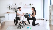 Concept of rehabilitation of disabled people. Young physiotherapist or wife in sportswear helping male patient in wheelchair exercise at home. Handicapped mature man training with stretching bands .