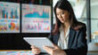 © pisan thailand - Asian business woman reviewing data in financial charts and graphs with tablet, in a modern office setting, focusing intently on her work, surrounded by digital screens displaying live market trends