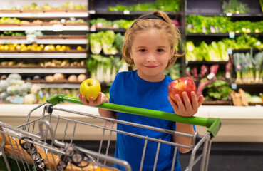 Wall Mural - Kid with apple and shopping cart at grocery store. Funny cute child on shopping in supermarket. Grocery store. Grocery shopping, healthy lifestyle concept.