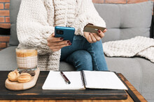Unrecognizable Woman Sitting On A Couch, Holding Credit Card And Using Mobile Phone Or Smartphone. Glass Of Coffee Drink With Dessert And Notepad With A Pen On The Table