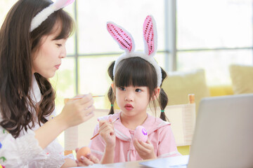 Asian young happy mother and little cute children daughter girl wearing funny bunny ears headbands with painting eggs while sitting together on table have laptop computer. Family celebrating Easter.