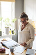 © Maskot - Mature woman photographing financial bills while standing in kitchen at home