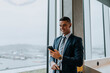 © Maskot - Smiling young businessman with smart phone standing by window at office
