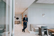 © Maskot - Full length of smiling young businessman with smart phone leaning on wall at corporate office