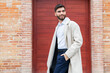 © ADDICTIVE STOCK - A stylish young man in business attire smiles confidently while standing against a red door backdrop, exuding a sense of casual professionalism in his beige coat and navy trousers