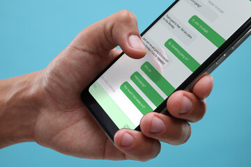 Man texting with friend using messaging application on smartphone against light blue background, closeup
