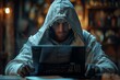 © Raptecstudio - A man in a hoodie sits in darkness at a table, using a laptop computer hacker