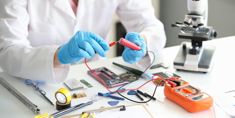 Canvas Print - A man with a voltmeter and a red battery in his hands, close-up. Tools and parts for building a device