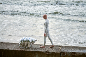 Young hairless girl with alopecia in white futuristic suit walking on concrete fence with toy tardigrade on sea background, bald pretty girl symbolizes courage and acceptance of unique appearance