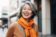 © pkproject - An elegant senior Asian woman with a stylish scarf smiles while walking in a vibrant city setting.