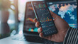© MP Studio - close-up of a hand holding a smartphone with stock market data on the screen, with a laptop in the background displaying additional financial charts