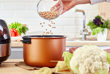 Female Hand Pours Peas Into A Multi Cooker. Multi-cooker Among Of Various Raw Foods On Kitchen Table. Woman Preparing Seed Or Cereal In A Slow Cooker.