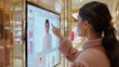 © weerasak - A young woman in a pink coat uses an interactive digital display to browse through fashion items in a luxury store.