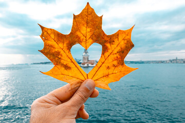 Wall Mural - Photo of the Maiden's Tower on the Bosphorus. The man is holding the leaf in his hand. Photo of the tower with an autumn leaf cut into a heart shape. The tower is blurred in the photo.
