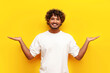 © Богдан Маліцький - cheerful curly indian man in white t-shirt holds empty hands to sides on yellow isolated background, young guy raises his hands and holds two options and smiles