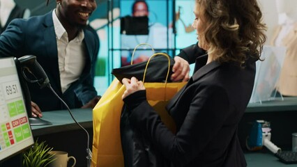 Canvas Print - Retail clerk putting clothes in a paper bag for customer, working at cash register and selling formal fashion items at department store. Woman employee assisting african american client. Camera A.