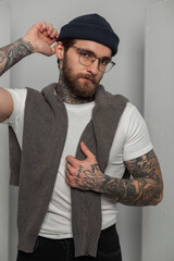 Wall Mural - Cool stylish young hipster man with a beard and tattoos with fashion glasses in a white T-shirt with a sweater puts on a knitted hat on a white background