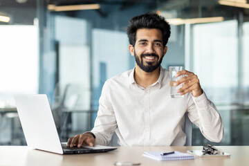 An Indian young male businessman is sitting in the office at his desk and holding a glass of clean water in his hand. He looks at the camera with a smile