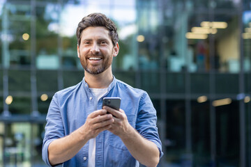 Close-up portrait of a young smiling man standing outside an office center on the street, holding a phone and looking at the camera
