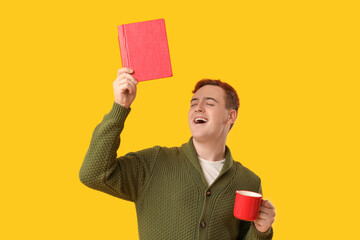 Wall Mural - Emotional young man with book and cup of hot beverage on yellow background