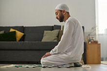 Young Muslim Man In White Skullcap And Thobe Standing On Knees On Rug And Performing Namaz Prayer Against Couch With Cushions