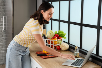 Poster - Young woman with fresh vegetables using laptop in kitchen