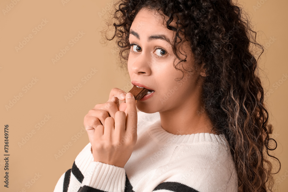 Beautiful young African-American woman eating piece of sweet chocolate on brown background