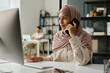 © pressmaster - Young serious female economist in hijab looking at desktop computer screen and talking to colleague or client on mobile phone