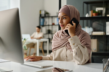 Leinwandbilder - Young serious female economist in hijab looking at desktop computer screen and talking to colleague or client on mobile phone