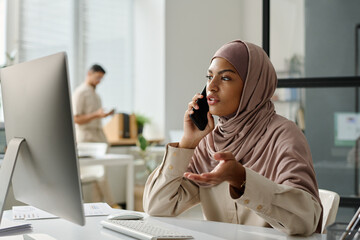 Leinwandbilder - Young Muslim female employee in hijab explaining something to business partner on phone while sitting by workplace with desktop computer