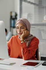 Leinwandbilder - Young bored businesswoman in hijab sitting in front of desktop computer and looking at screen while waiting for page uploading