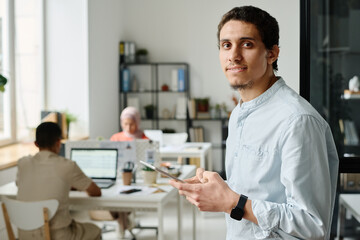 Leinwandbilder - Young Muslim male entrepreneur or office manager with tablet looking at camera with smile while standing against colleagues in coworking space