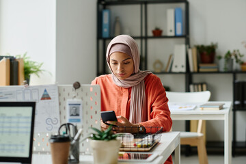 Leinwandbilder - Young Muslim female office worker in hijab sitting by workplace in coworking space and scrolling through posts in smartphone